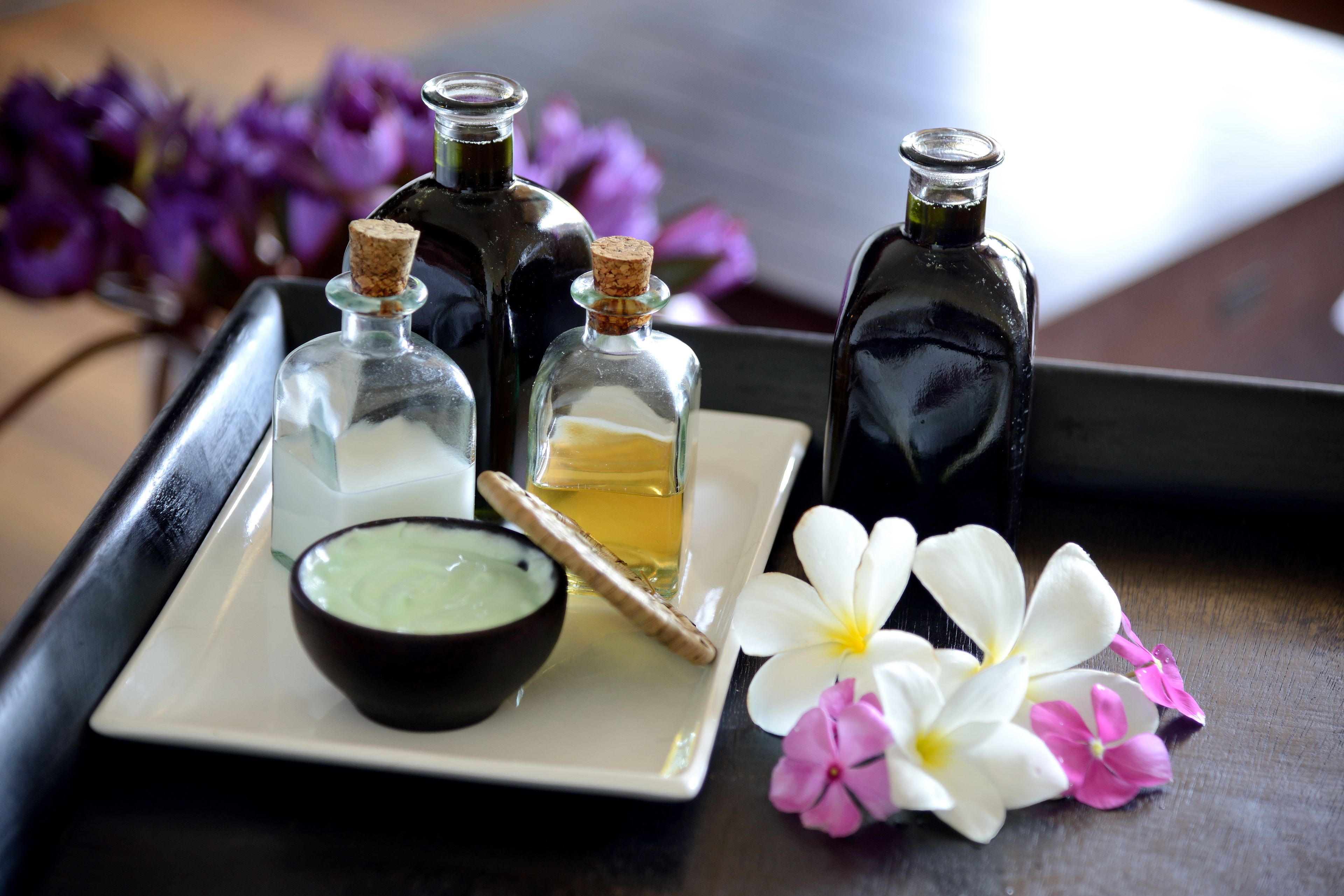 A tray with three glass bottles, a bowl of green substance, a wooden stick, and two flowers.