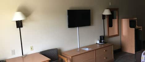 A hotel room with a wooden desk, a chair, a television mounted on the wall, a bedside table with a lamp, and a microwave.