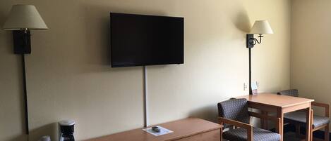 A hotel room with a TV mounted on the wall, a wooden dresser with drawers, a table with two chairs, and two floor lamps.