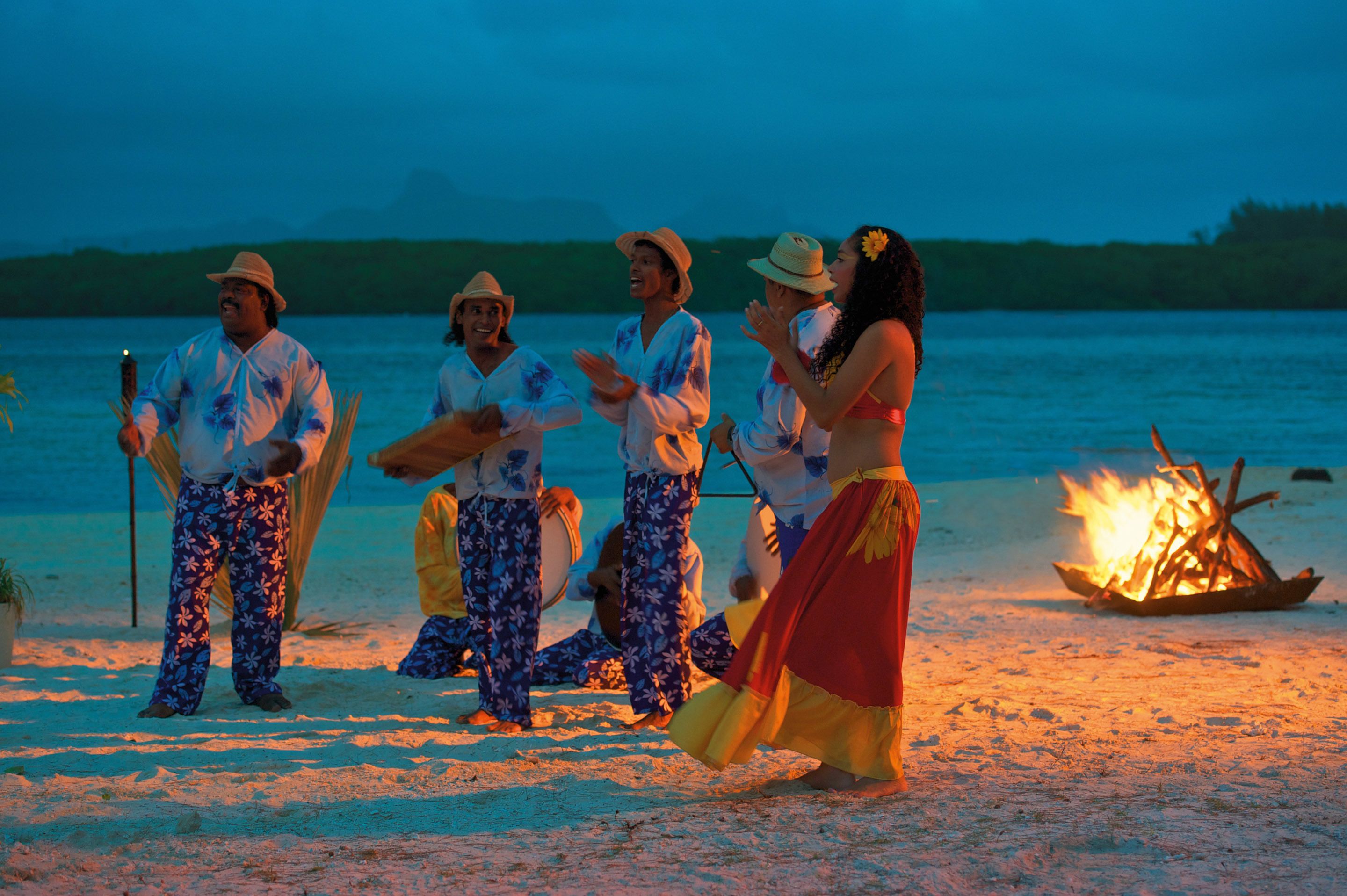 A group of people performing a traditional dance on a beach with a fire in the background.