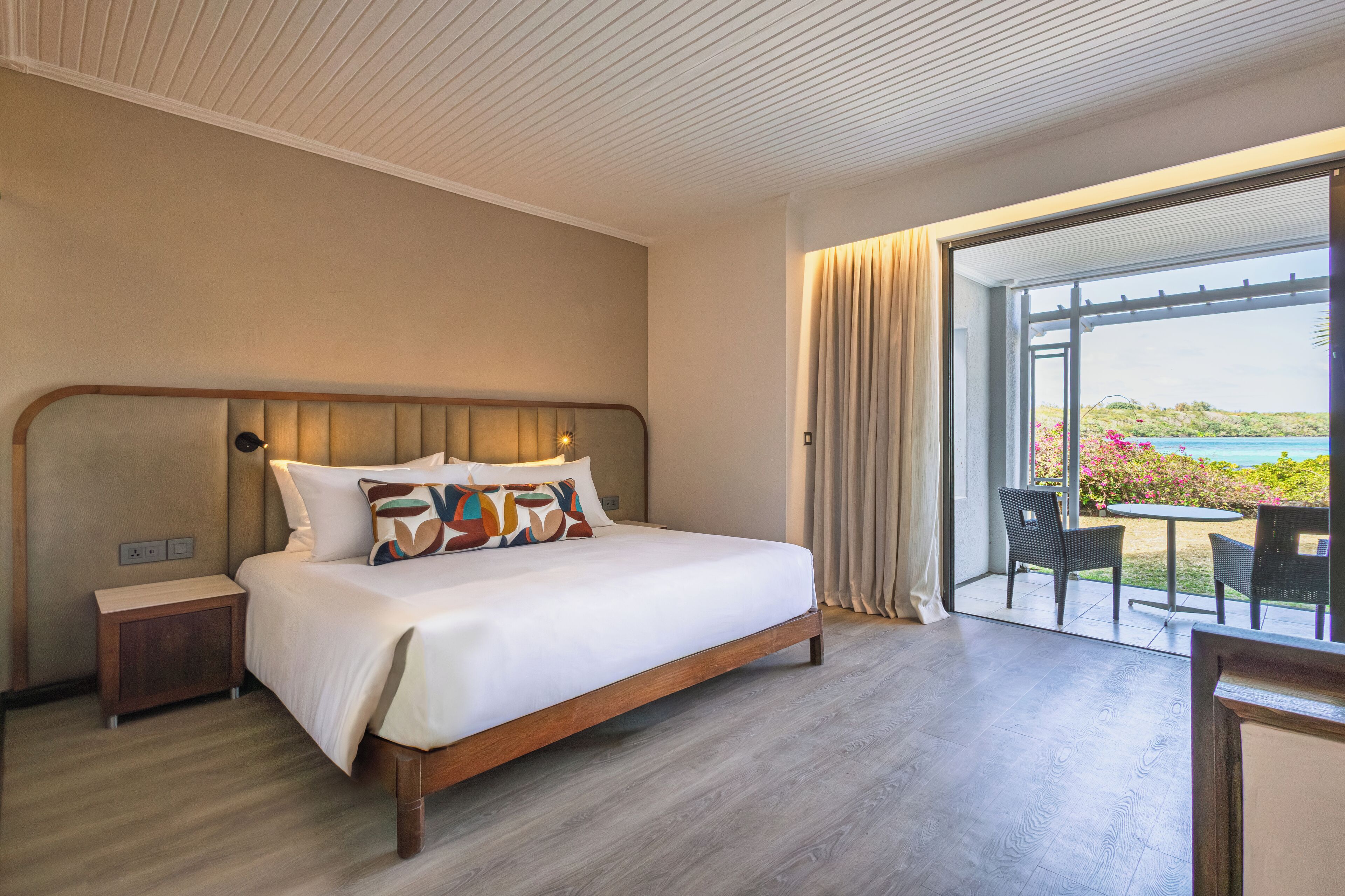 A modern hotel room with a large bed, a nightstand, and a view of the ocean through a sliding glass door.