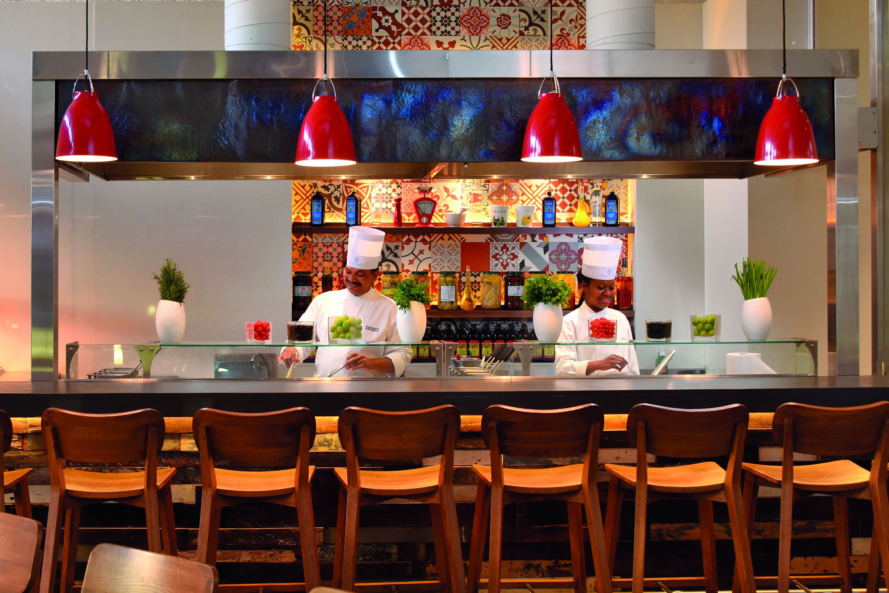 A modern kitchen with chefs preparing food, a bar area with wooden chairs, and a colorful backsplash.
