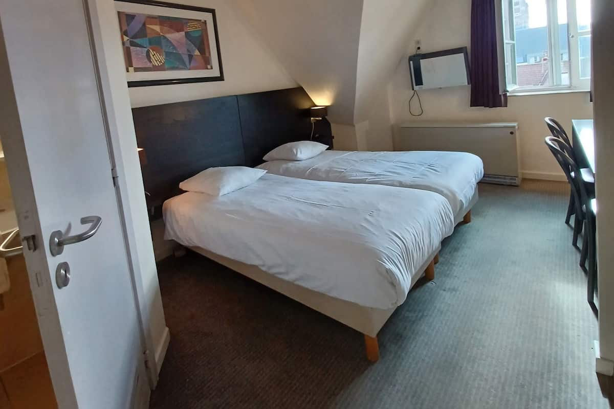 Une chambre d’hôtel avec deux lits, un bureau, une fenêtre avec des rideaux, un tableau au mur et un luminaire au plafond.