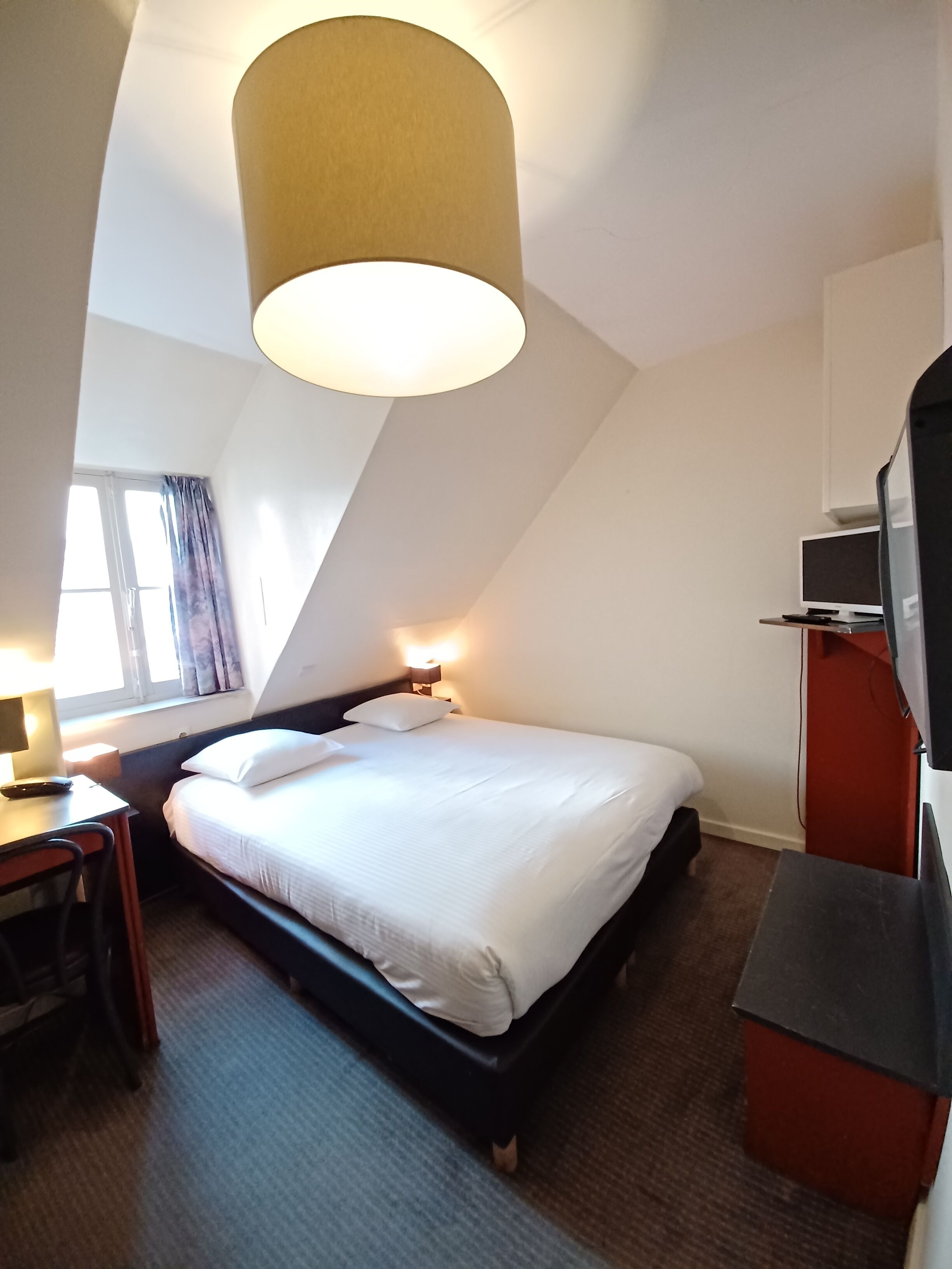 Ein Hotelzimmer mit Bett, Schreibtisch, Lampe, Fernseher und einem Fenster mit Vorhängen.