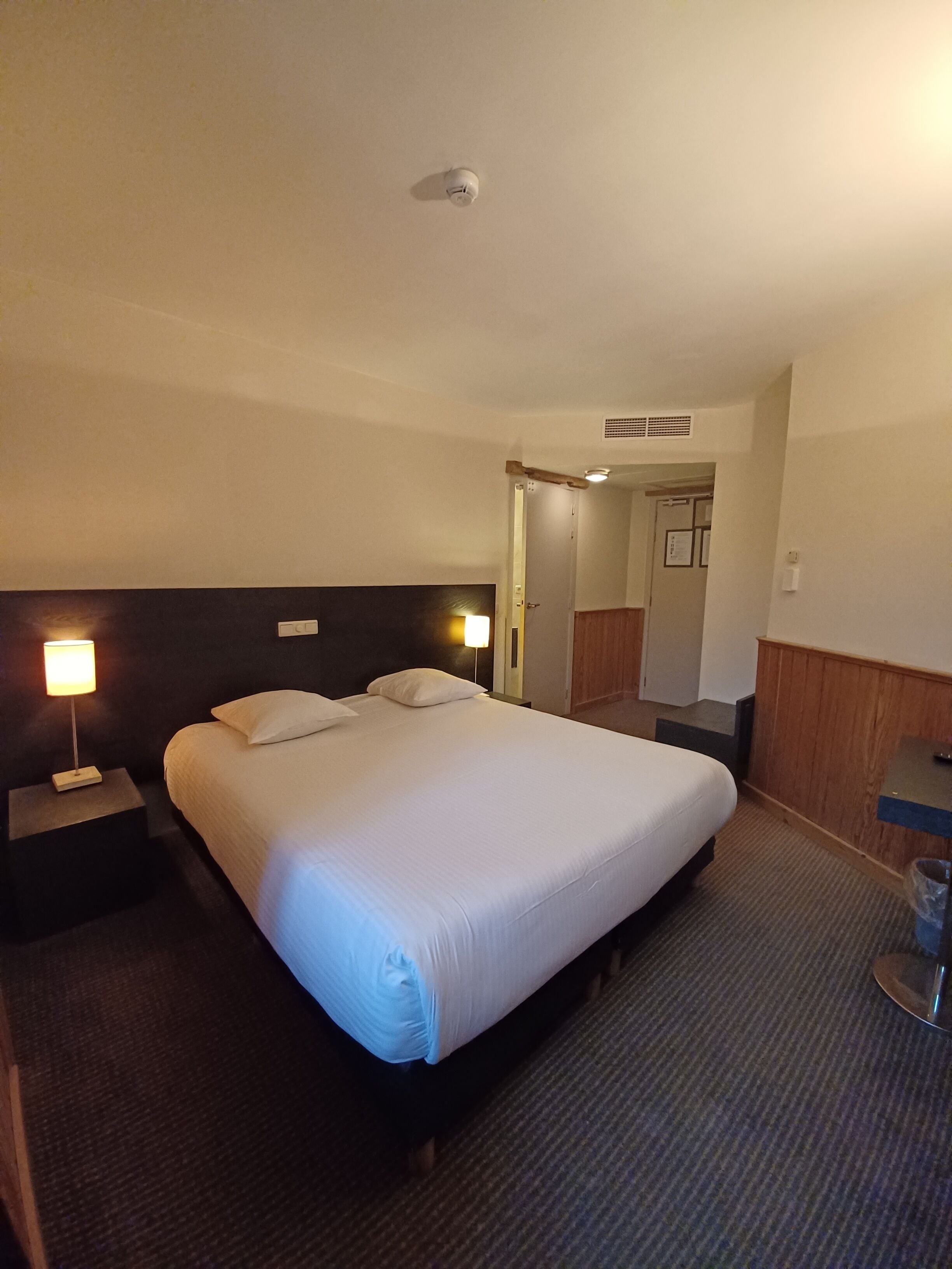A hotel room with a large bed, two bedside lamps, a wooden headboard, a small table, and a door leading to another room.