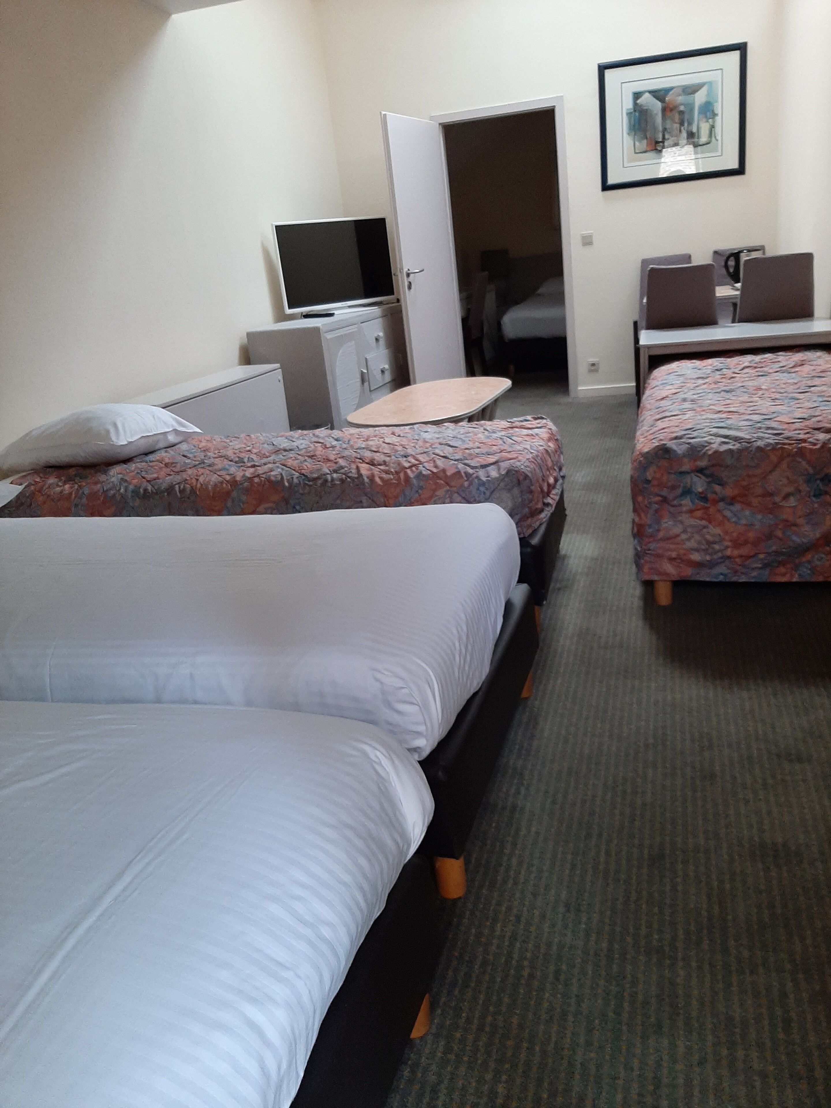 Habitación de hotel con dos camas, un escritorio, una silla, un televisor y una puerta que da a otra habitación.
