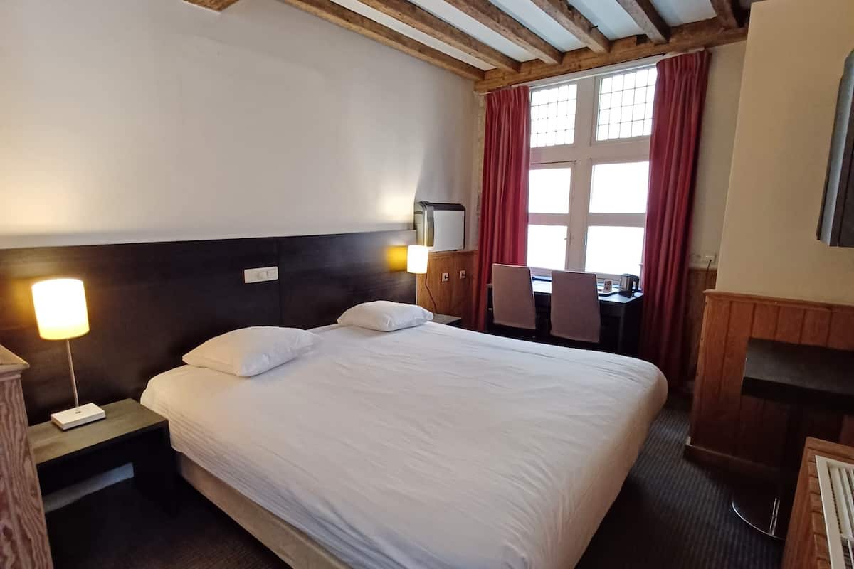 Une chambre d’hôtel avec un grand lit, des poutres en bois au plafond, un bureau avec une chaise et une fenêtre avec des rideaux rouges.