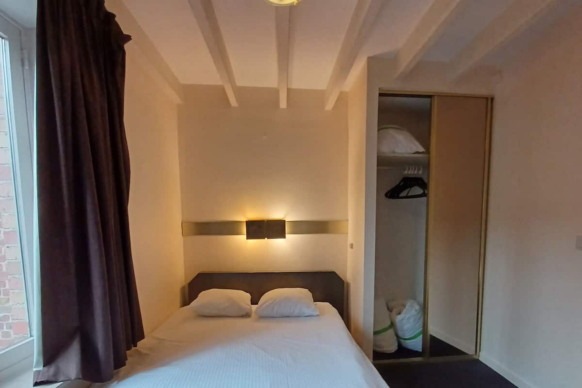 Une chambre d’hôtel avec un lit, une fenêtre avec des rideaux, un luminaire fixé au mur et une armoire contenant des vêtements suspendus.