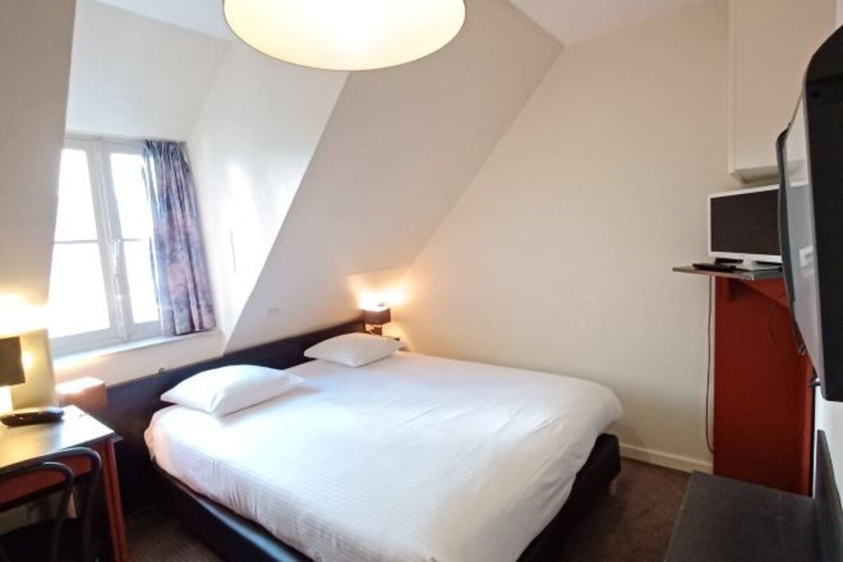 Une chambre d’hôtel équipée d’un lit, d’une table de chevet, d’une lampe, d’une télévision et d’une fenêtre avec des rideaux.