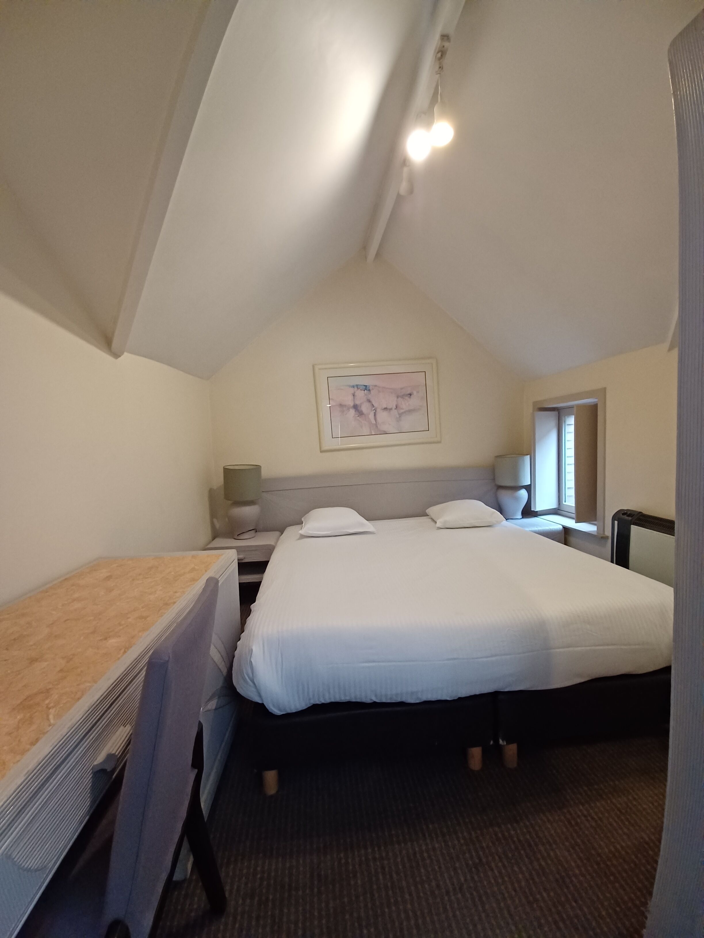 A bedroom with a bed, a desk, a chair, a nightstand, a lamp, a window, and a painting on the wall.
