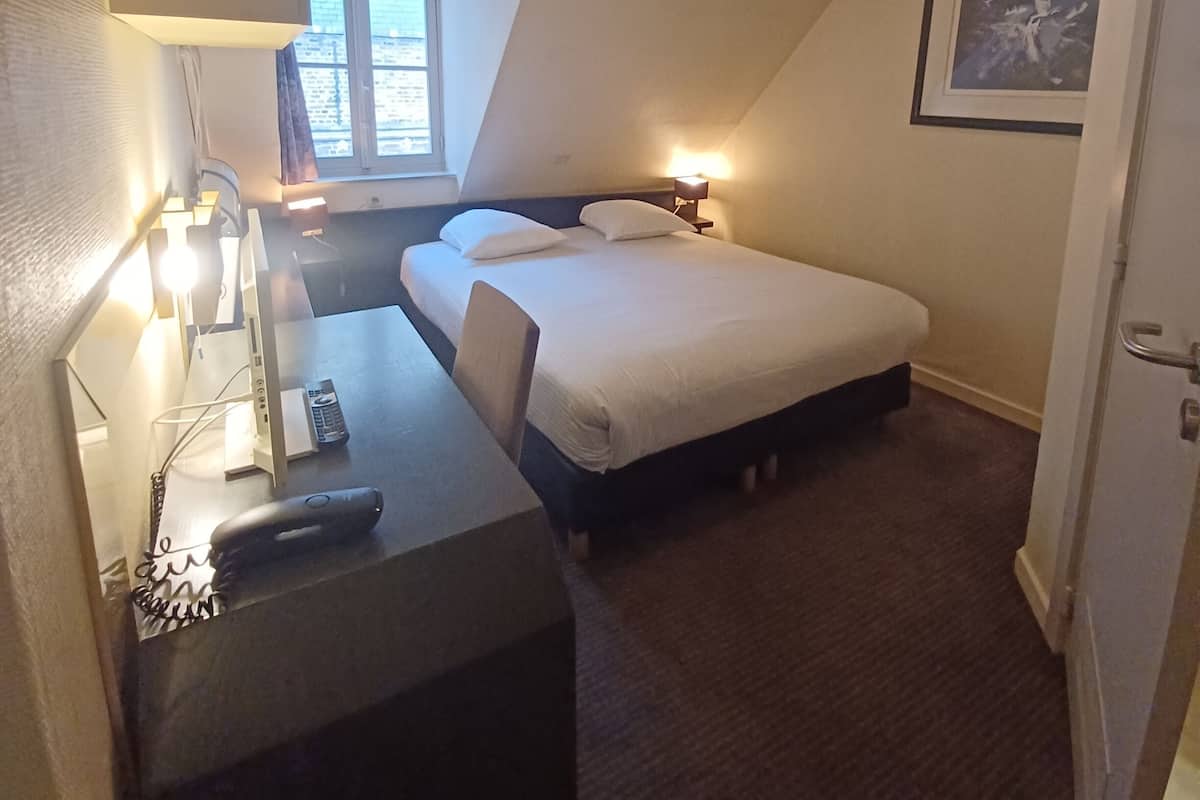 Une petite pièce faiblement éclairée, avec un lit, un bureau et une chaise. Il y a une fenêtre avec des rideaux, un tableau encadré au mur et une porte.