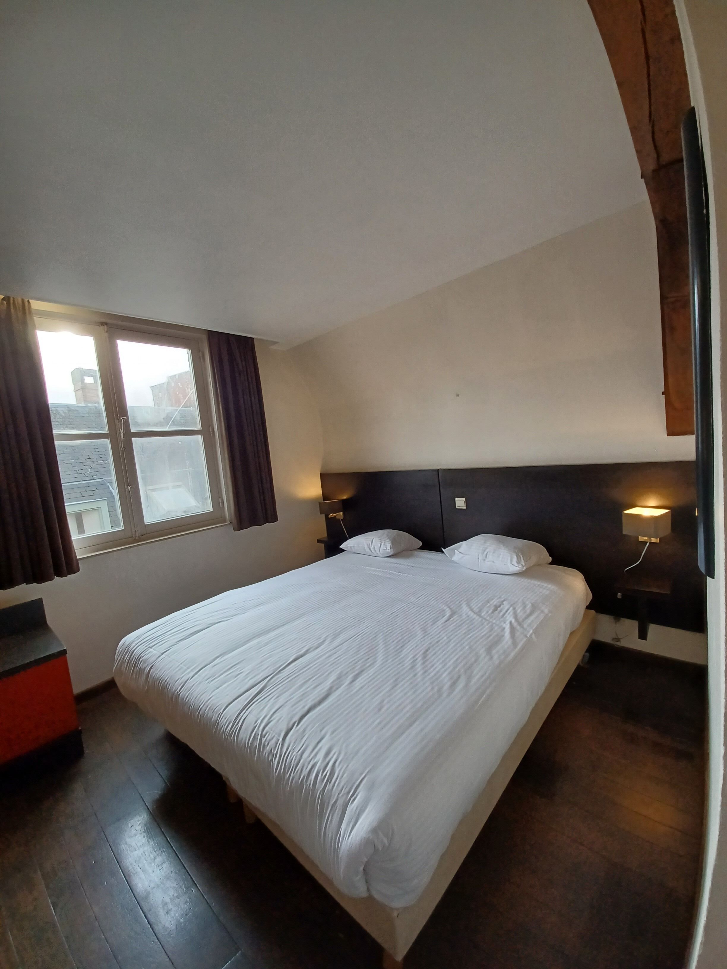 Une chambre avec un grand lit, une fenêtre avec des rideaux, une table de chevet avec une lampe et un plancher en bois.