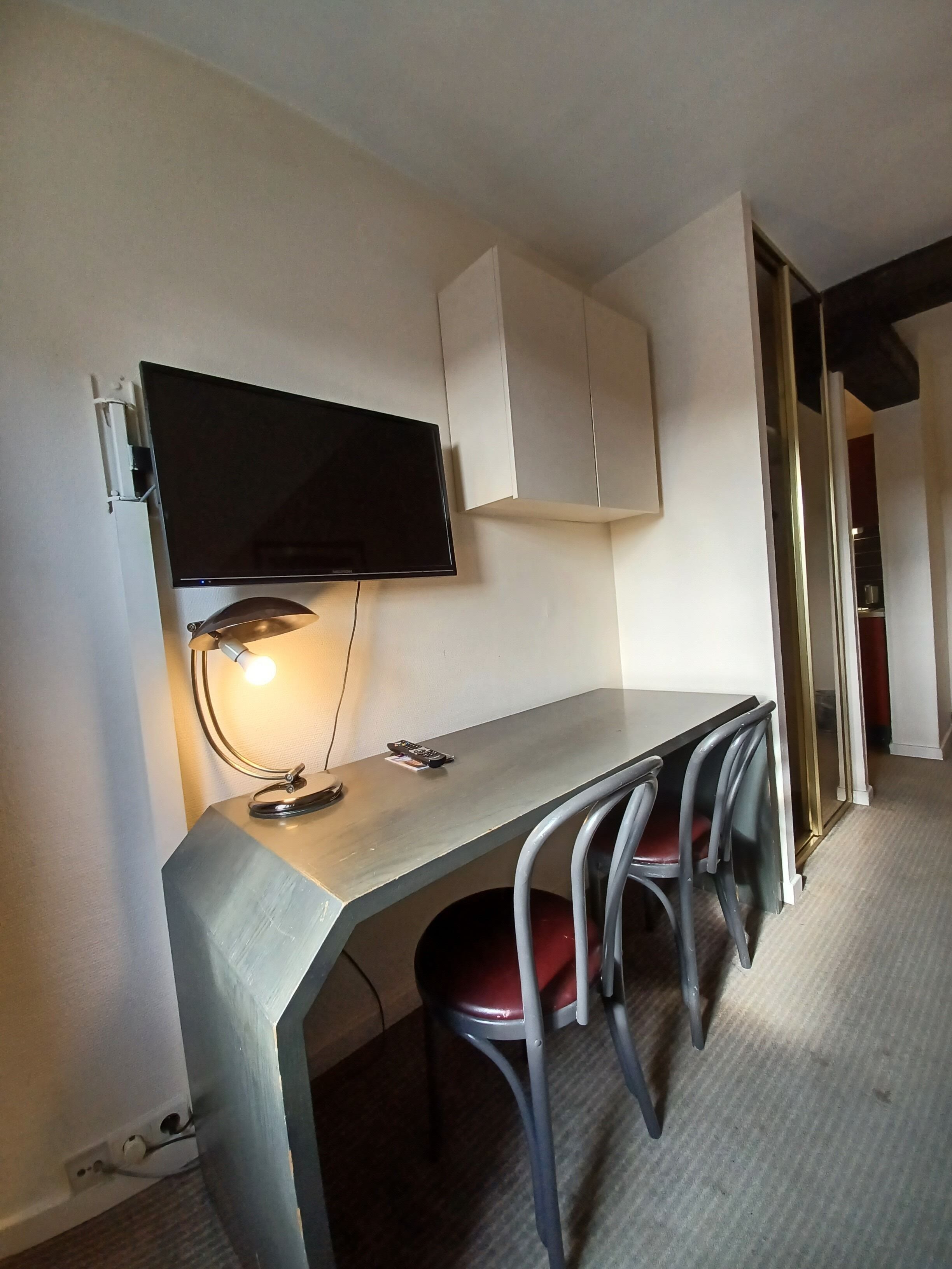 A compact room with a desk, a chair, a television mounted on the wall, and a cabinet.