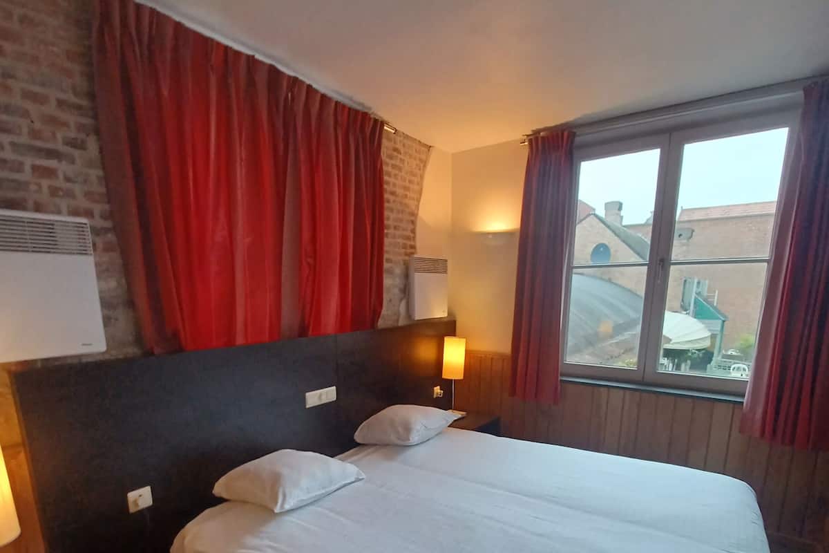 Une chambre d’hôtel avec un grand lit, une tête de lit, une table de chevet, une lampe et une fenêtre avec des rideaux.