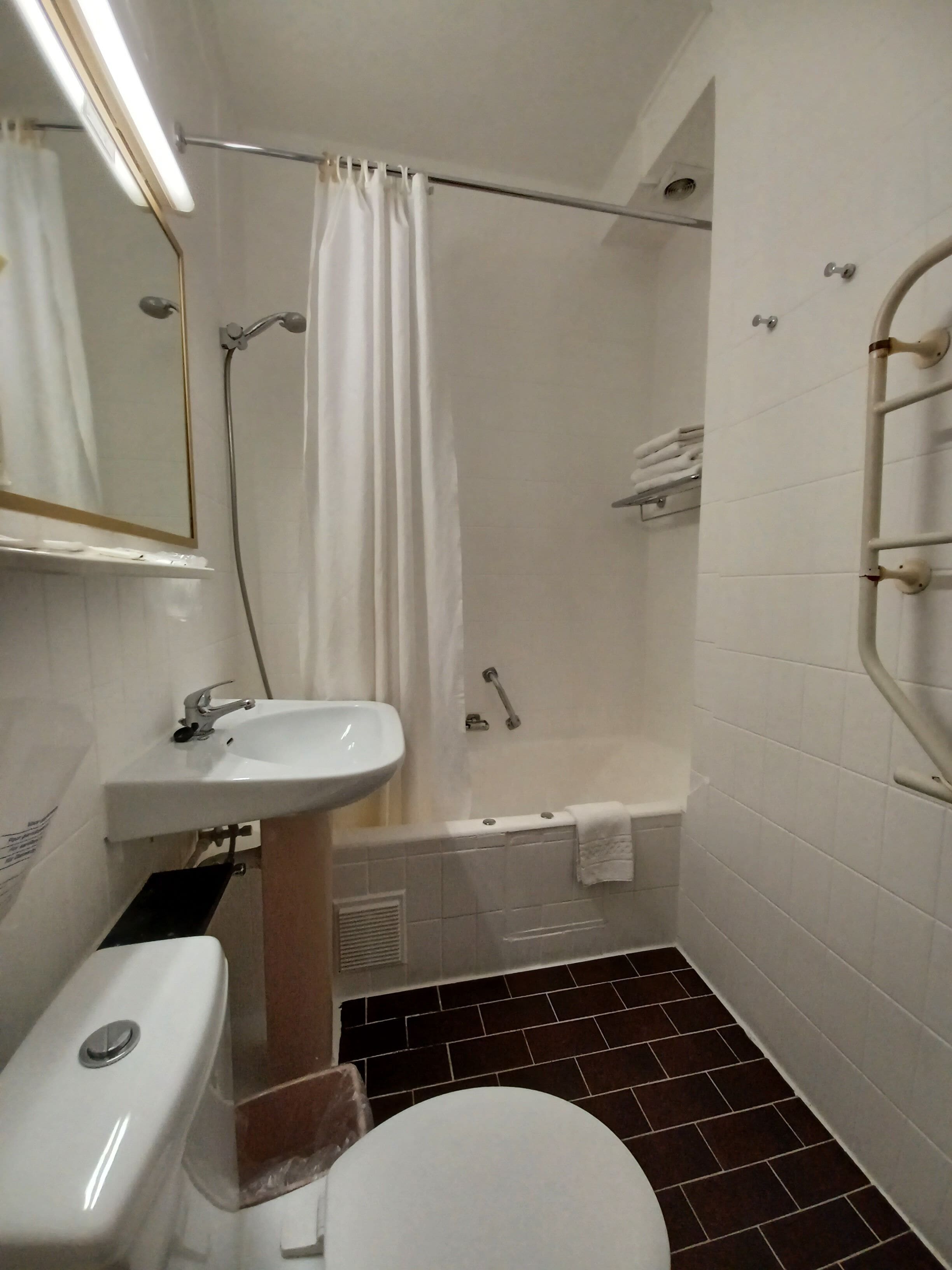 Un baño pequeño y funcional con lavamanos, inodoro y bañera blancos. Hay una cortina de ducha, un perchero para toallas y un pasamanos en la pared.