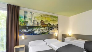 Habitación de hotel con dos camas, un cuadro de un paisaje urbano en la pared, una ventana con cortinas y una mesita de noche pequeña.
