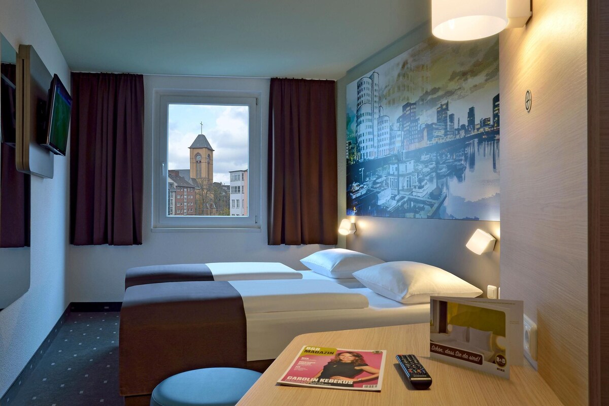 Ein Hotelzimmer mit einem Bett, einem Schreibtisch mit einer Zeitschrift, einer Fernbedienung und einem Fenster mit Blick auf ein Gebäude.