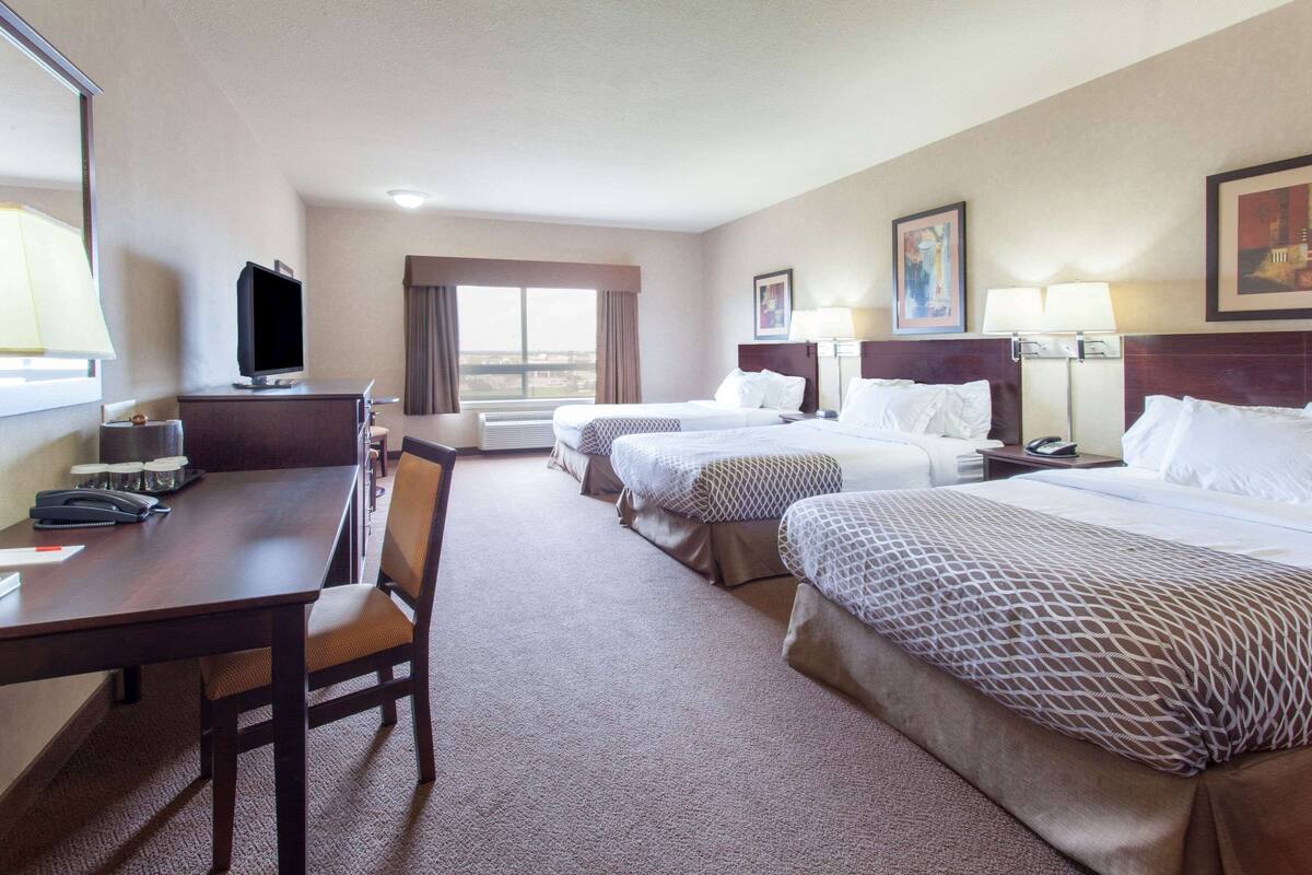Habitación de hotel con tres camas, un escritorio con silla, televisión y un ventanal grande.