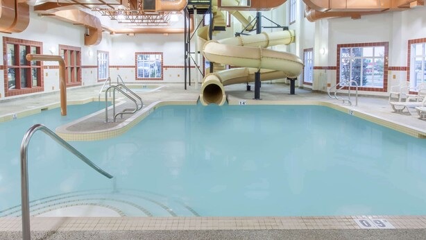 An indoor swimming pool with a slide, clear poolside area, and a view of the outside through large windows. - Ramada by Wyndham Camrose (Camrose)