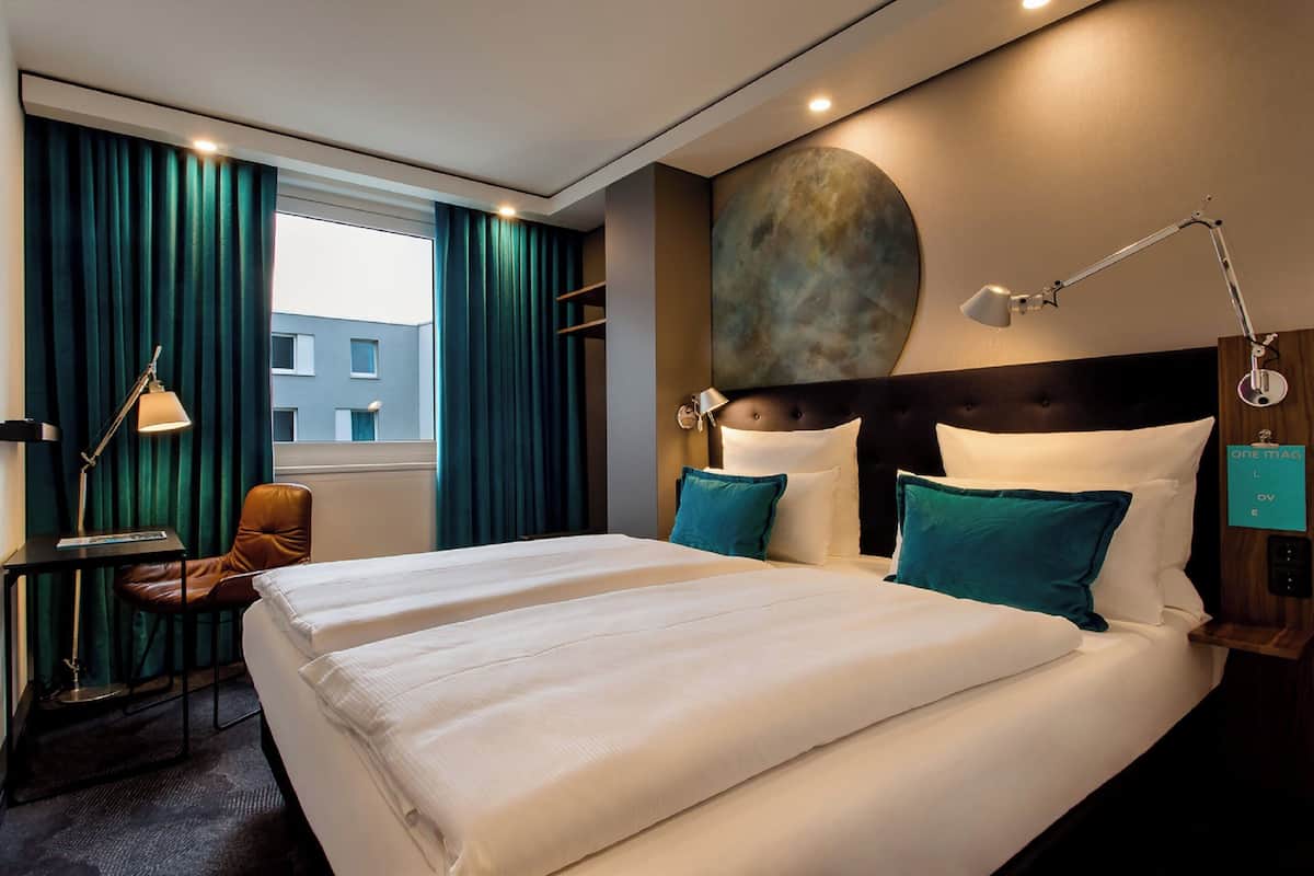 A hotel room with a large bed, two pillows, a desk with a chair, a lamp, and a view of the outside.