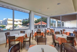 Un área de comedor interior con mesas y sillas, ventanas amplias que ofrecen vistas al exterior y una zona de piscina visible a través de las ventanas.