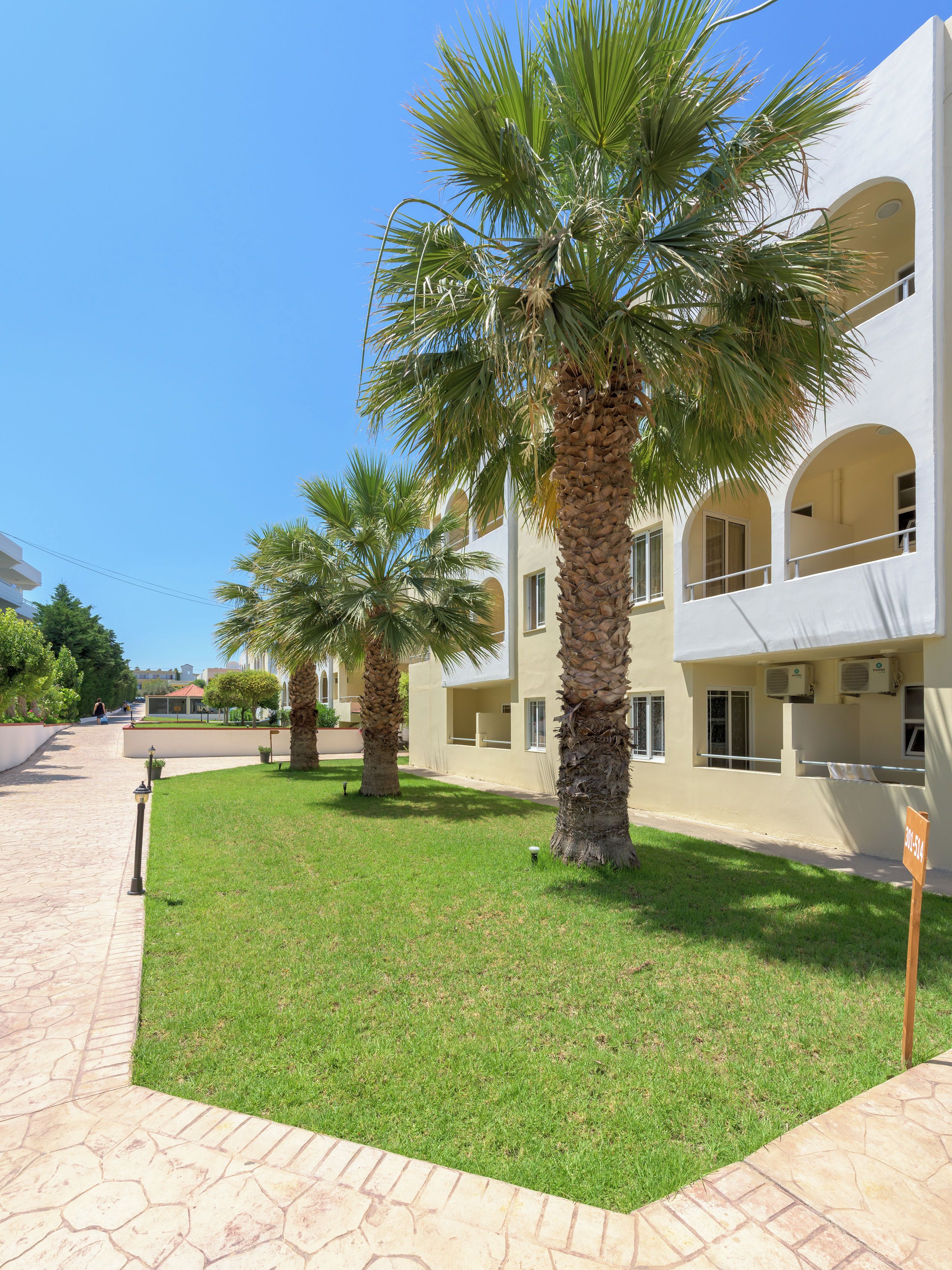Un patio con palmeras, un sendero empedrado y un edificio con balcones y ventanas.