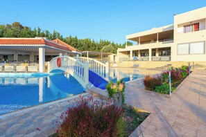 Una piscina con un puente, rodeada de jardines con plantas y un edificio con tejado de azulejos.