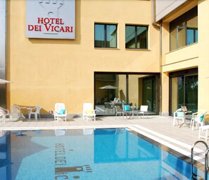 Exterior de un hotel con piscina, mobiliario exterior y un cartel que dice 'HOTEL DEI VICARI'.