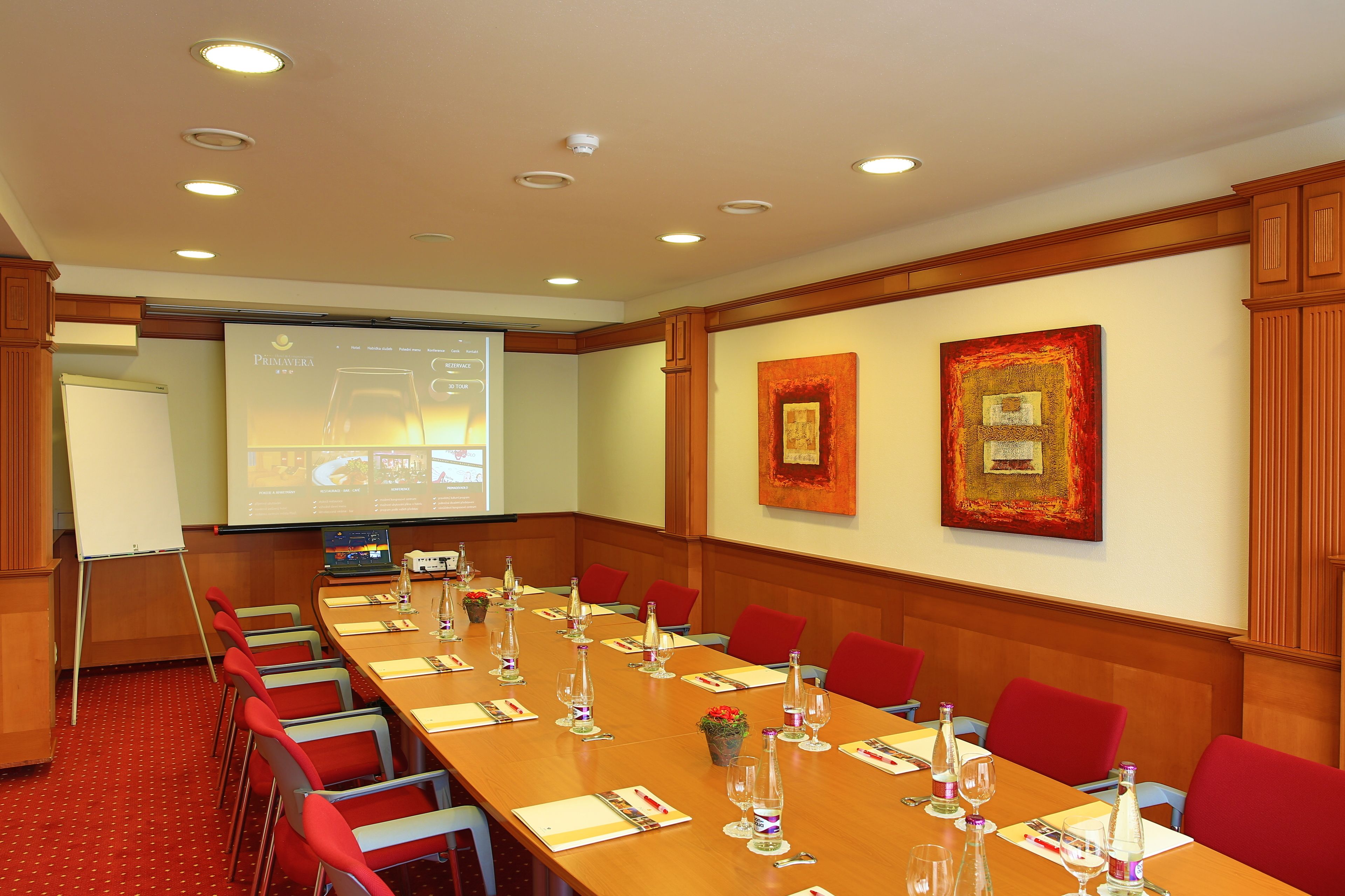 A conference room with a long table, red chairs, a projector screen, and framed artwork on the walls.