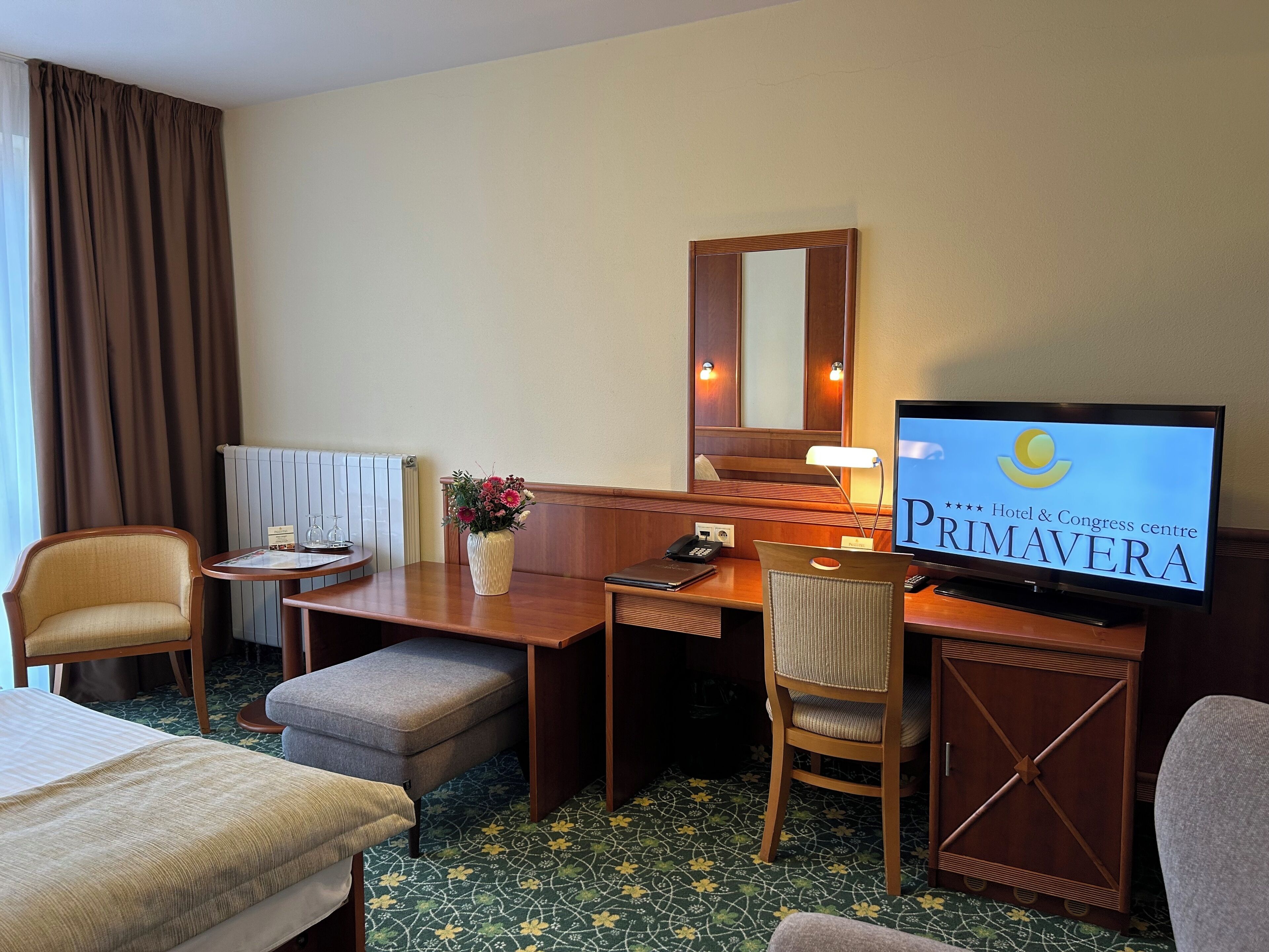 Ett hotellrum med en säng, ett skrivbord, en stol, en tv och ett fönster med gardiner.