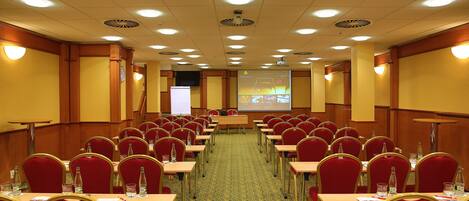 A conference room with rows of red chairs, tables, and a projector screen at the front.