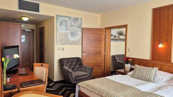 A hotel room with a bed, a desk with a computer and lamp, a grey armchair, a wooden wardrobe, and a painting on the wall.