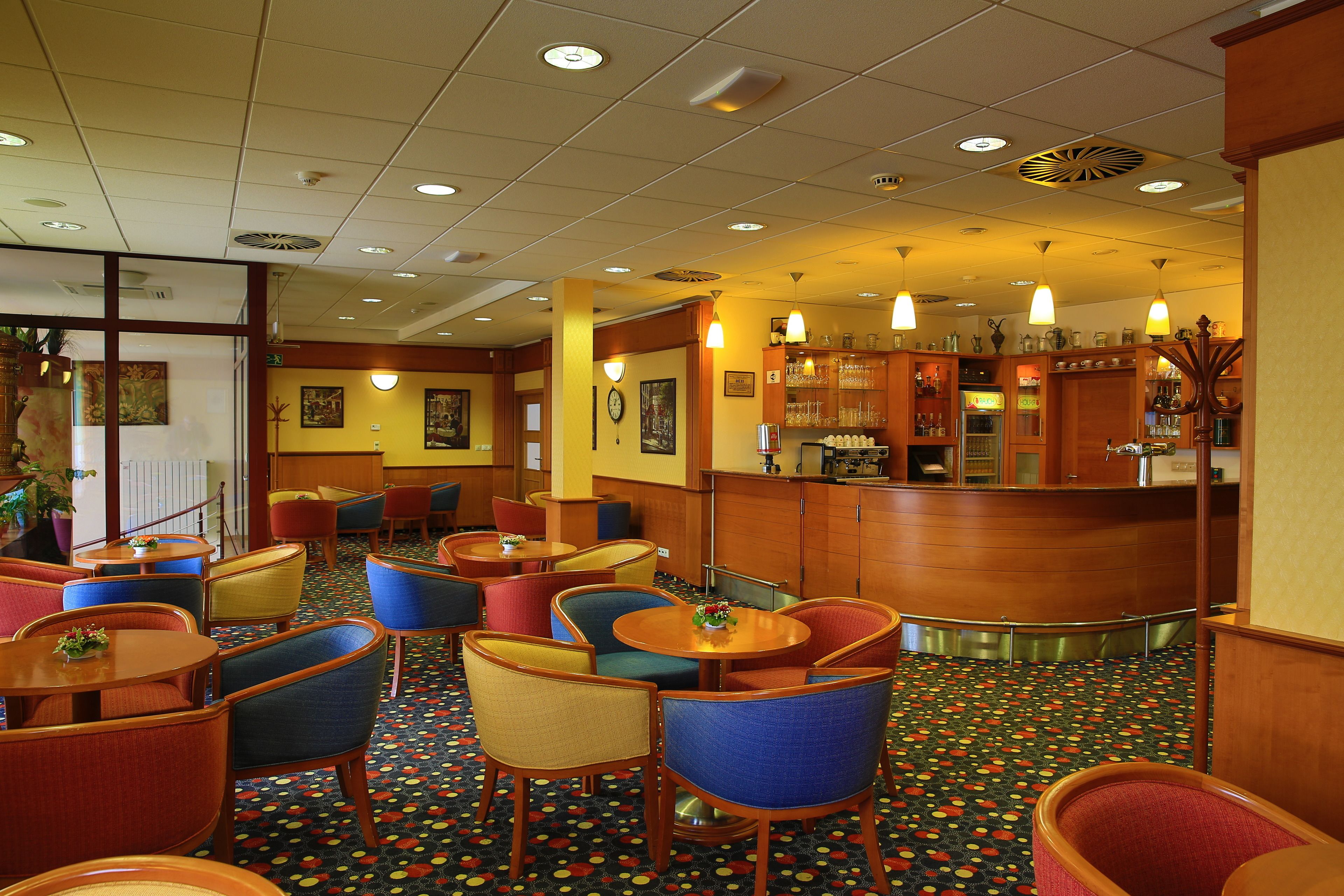 A hotel lobby with round tables, colorful chairs, a bar area, and a patterned carpet.