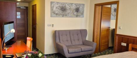 A hotel room with a wooden desk, a television, a gray armchair, a bed, and a floral arrangement.