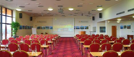 A conference room with rows of red chairs and tables, a projector screen, and a carpeted floor.