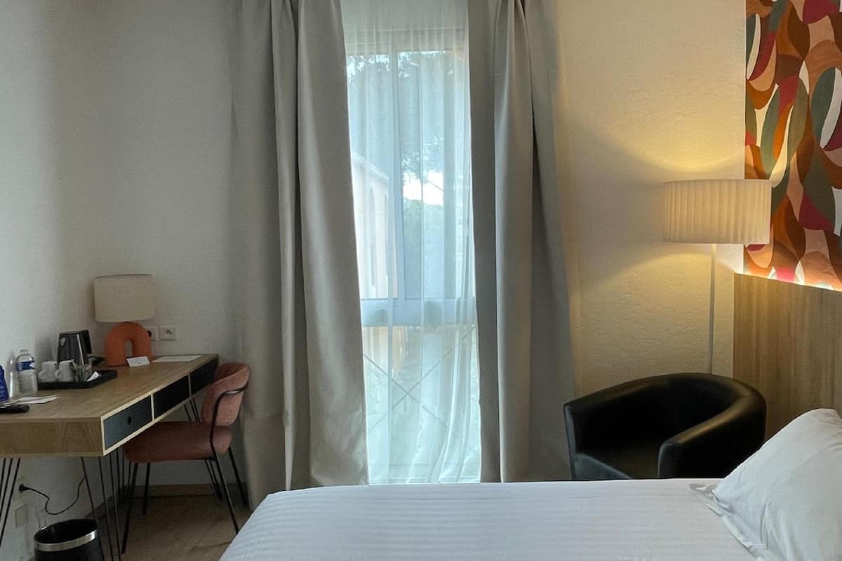 A hotel room with a bed, a desk with a chair, a lamp, and a window with curtains.