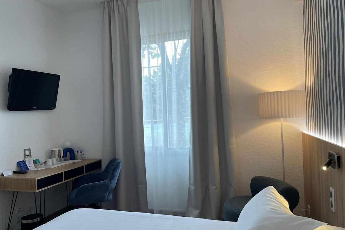 Habitación de hotel con cama, escritorio con silla, televisor y ventana con cortinas.