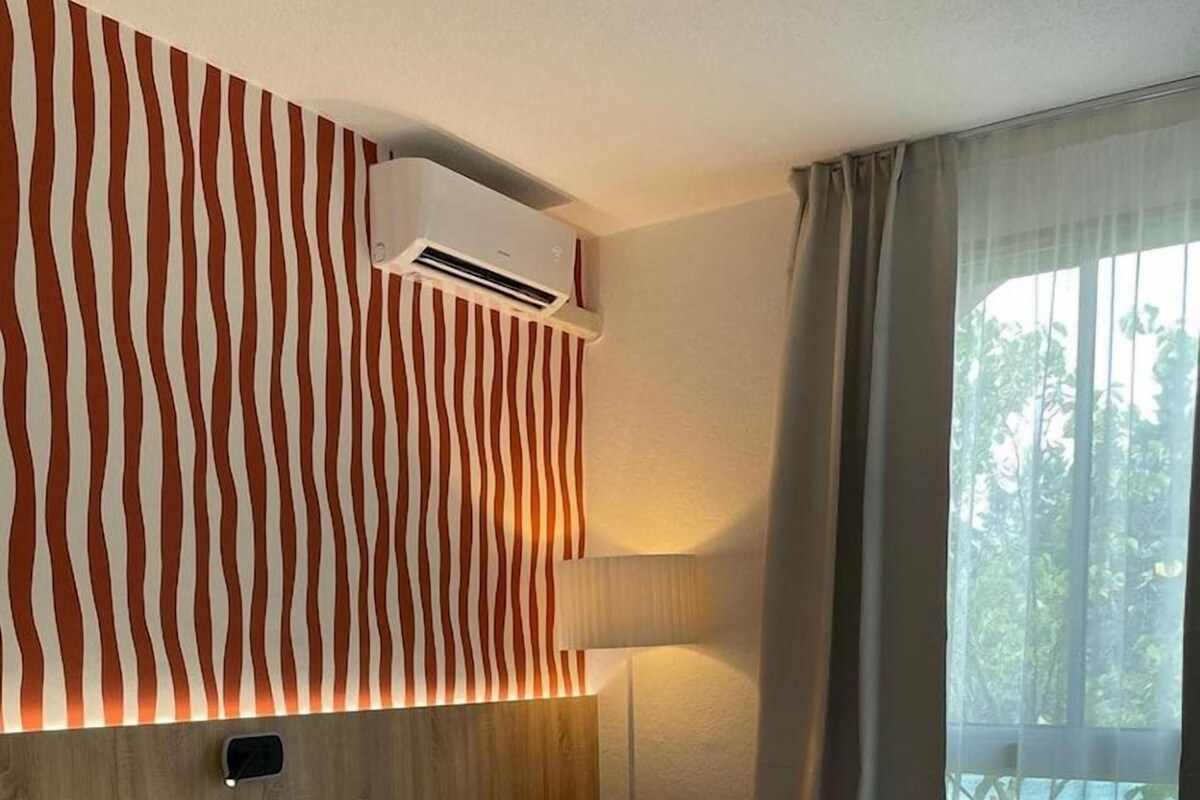 Habitación de hotel con una cama grande, una silla roja, un equipo de aire acondicionado y una ventana con cortinas.