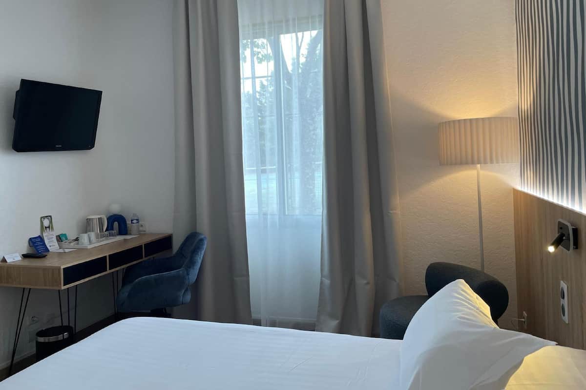 A hotel room with a bed, a desk with a chair, a television, and a window with curtains.