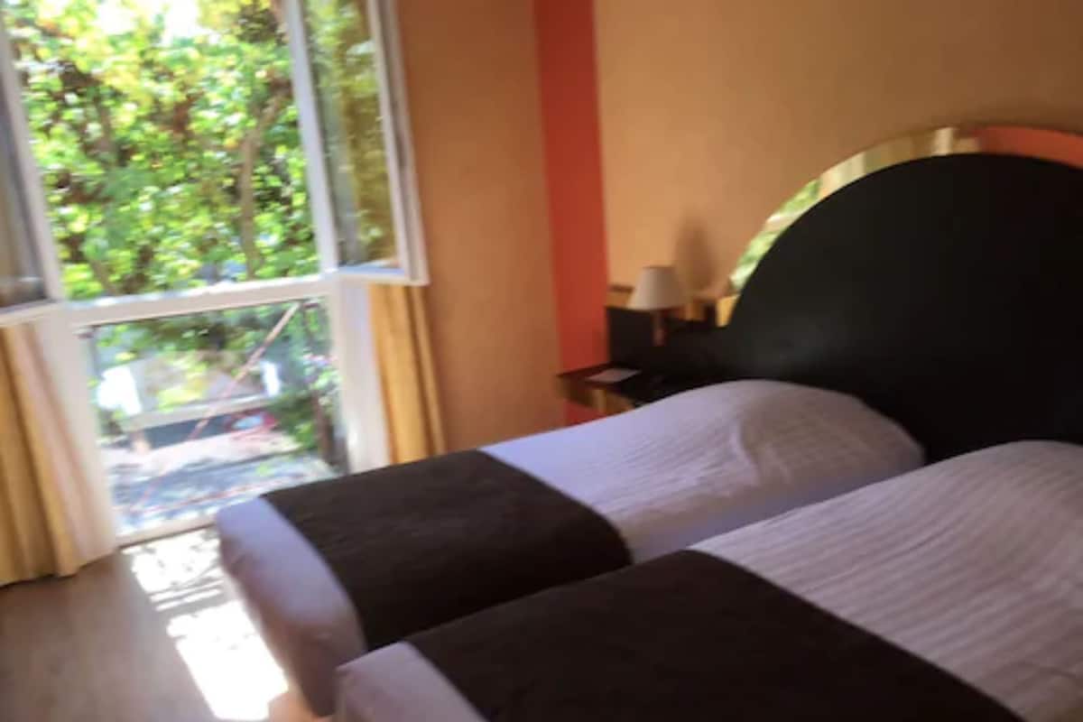 A hotel room with a bed, a nightstand, a window with curtains, and a view of greenery outside.
