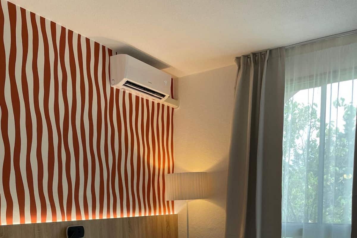 A hotel room with a large bed, a red chair, a wall-mounted air conditioner, and a view of greenery through the window.