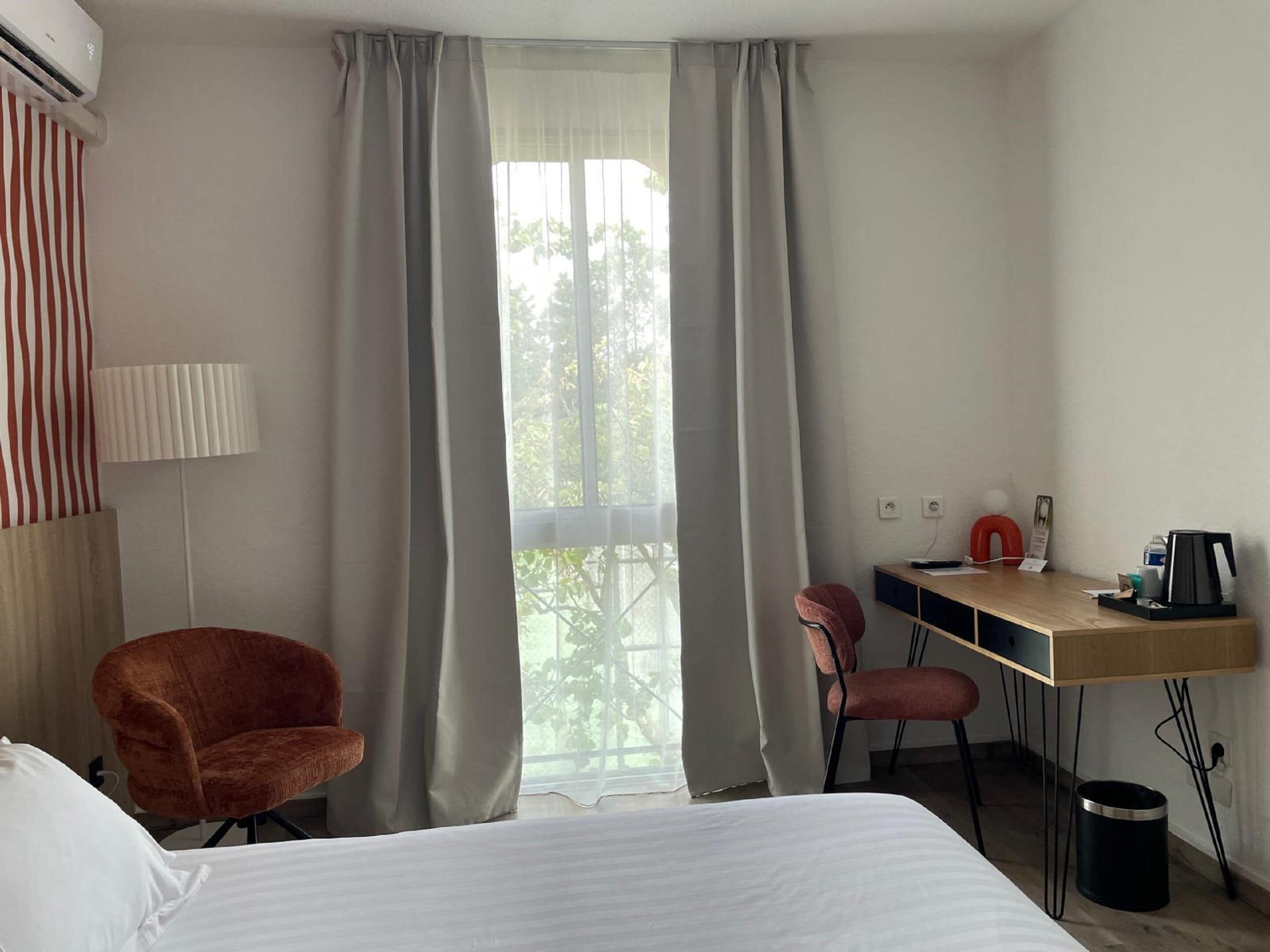 Ett hotellrum med en säng, ett skrivbord, två stolar, en lampa och ett fönster med gardiner.