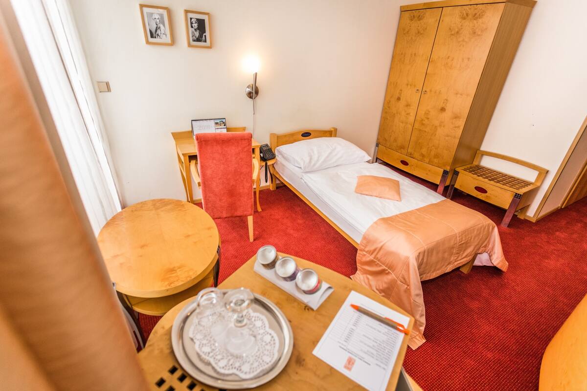 A hotel room with a single bed, a wooden wardrobe, a desk with a chair, a small table with a tray, and a red carpet.