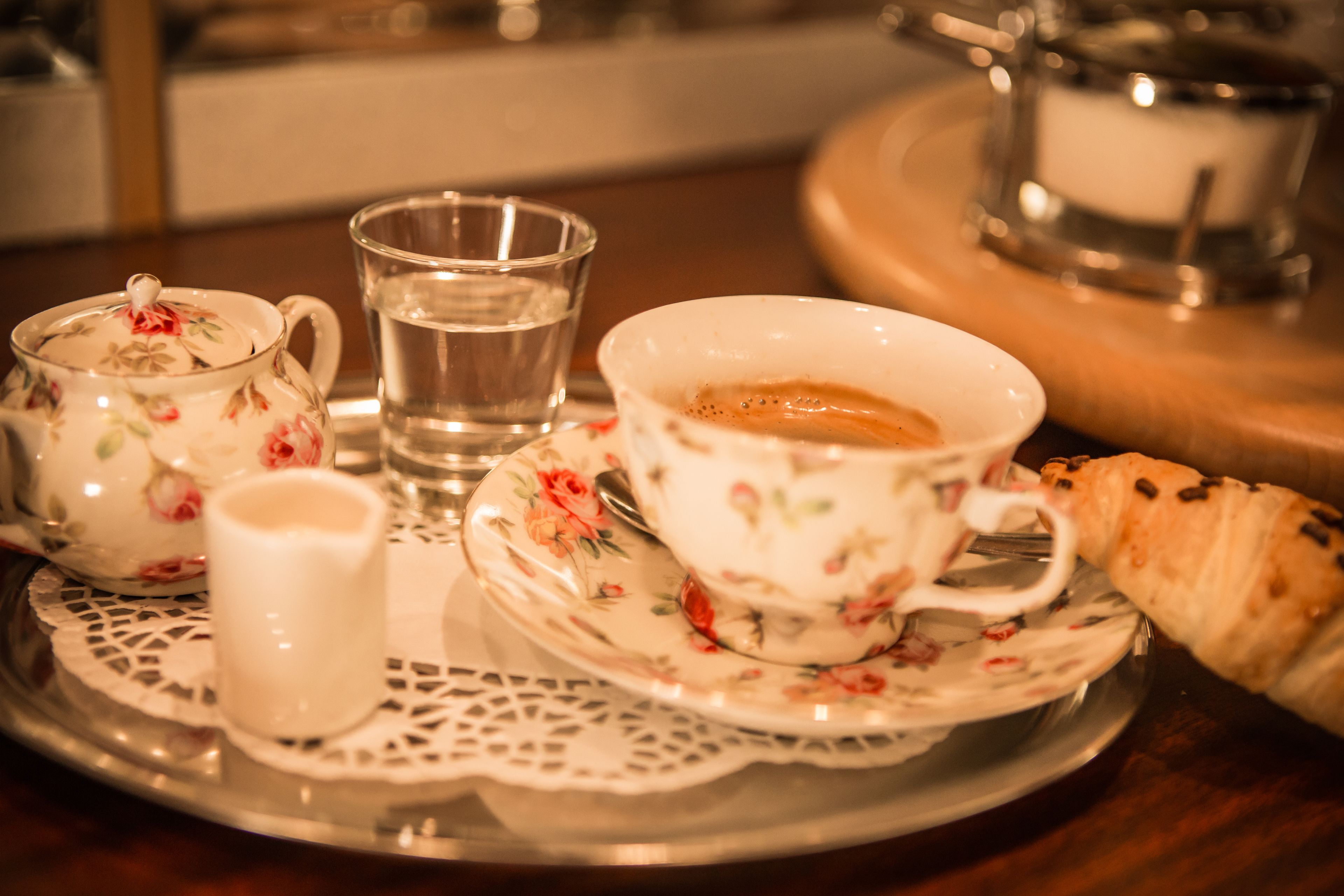 A tray with a floral teapot, a cup of coffee, a glass of water, a small cup, and a croissant.