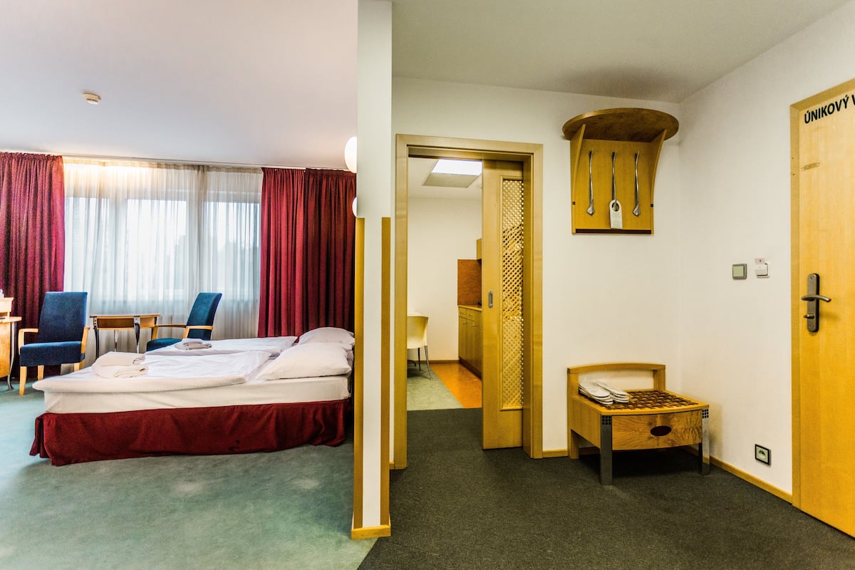 A hotel room with a bed, a desk, a chair, and a small table with a chair. There is a door with a wooden panel and a coat rack above it.
