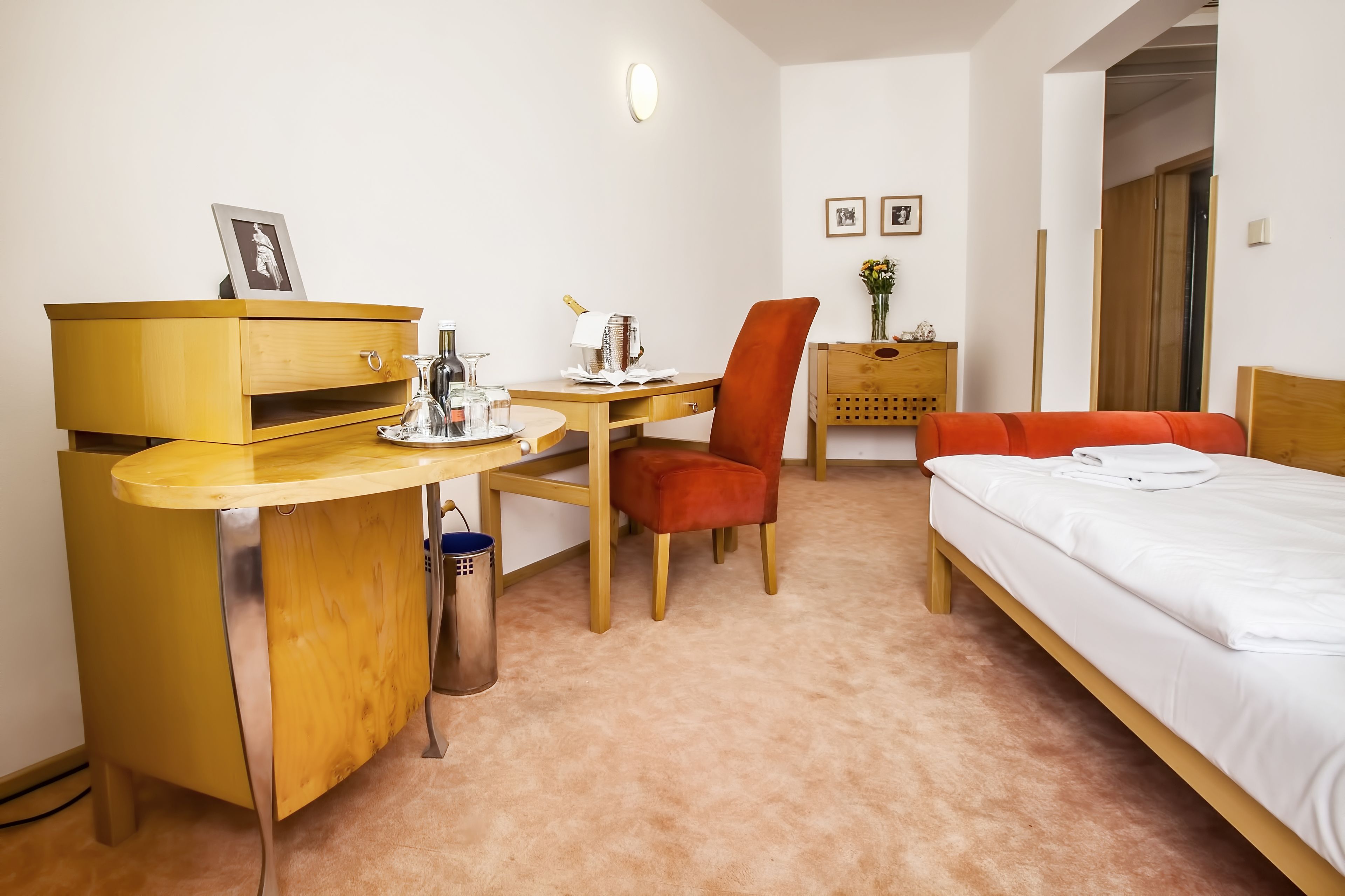 A hotel room with a wooden desk, a red chair, a bed with white bedding, and a small wooden nightstand with a vase of flowers.