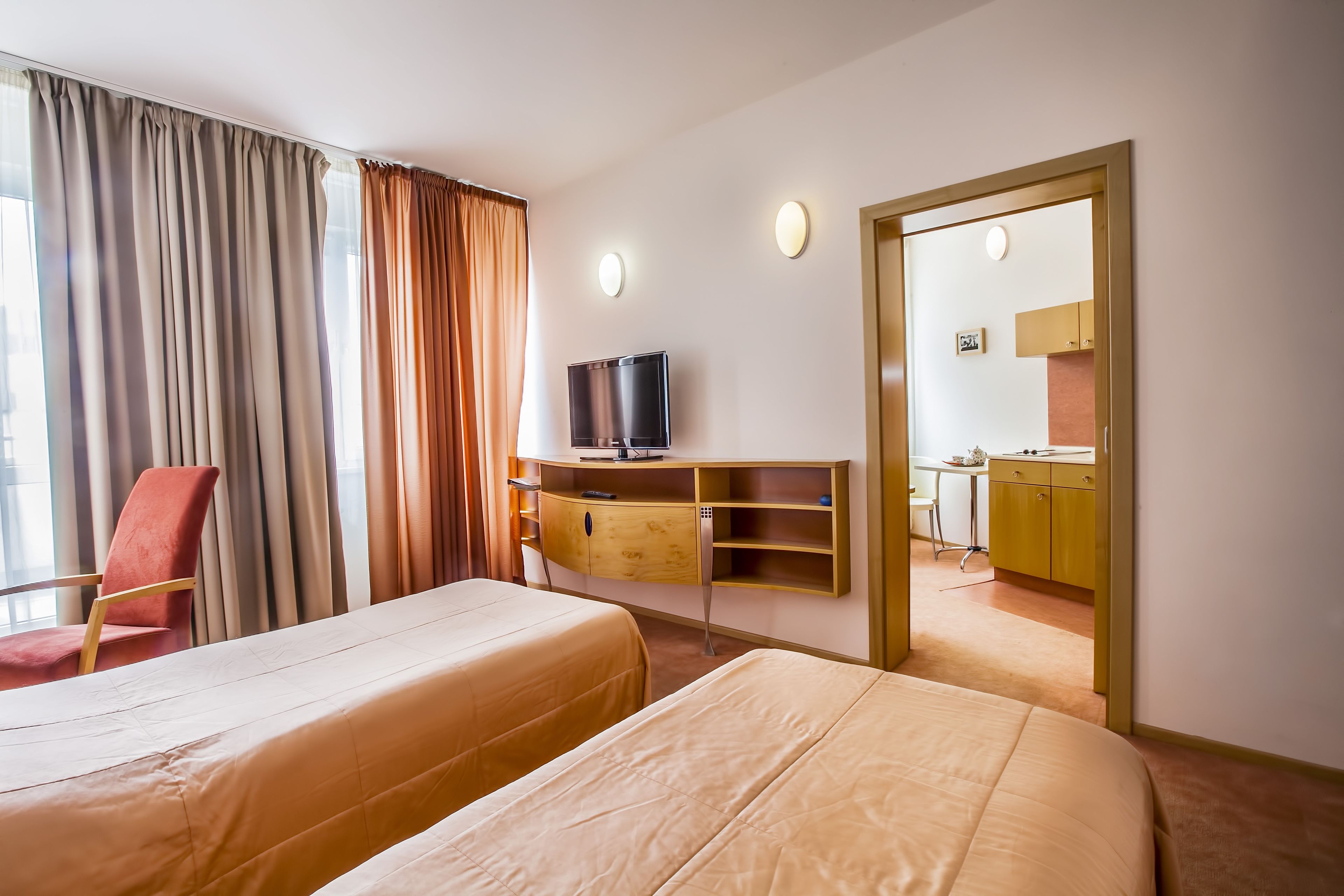 A hotel room with two beds, a TV, a desk, and a view of the kitchen.