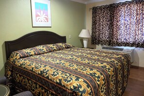 A bed with a patterned bedspread, a bedside lamp, a window with patterned curtains, and a framed picture on the wall.