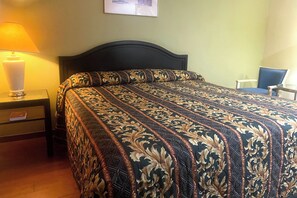 A neatly made bed with a patterned bedspread, a bedside table with a lamp, and a chair in a room with plain walls.