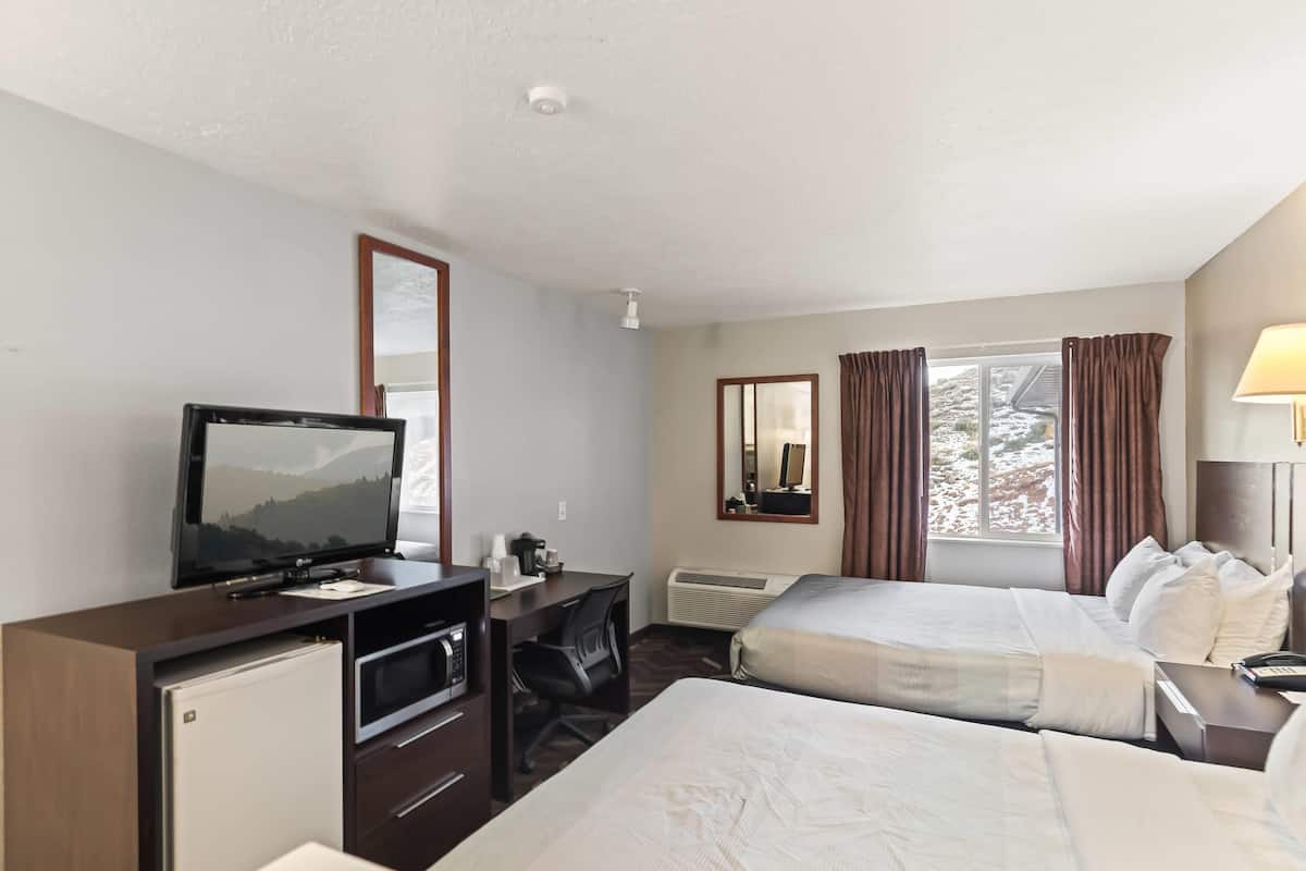 A hotel room with a bed, a desk with a television, a mini-fridge, a chair, a window with curtains, and a lamp.